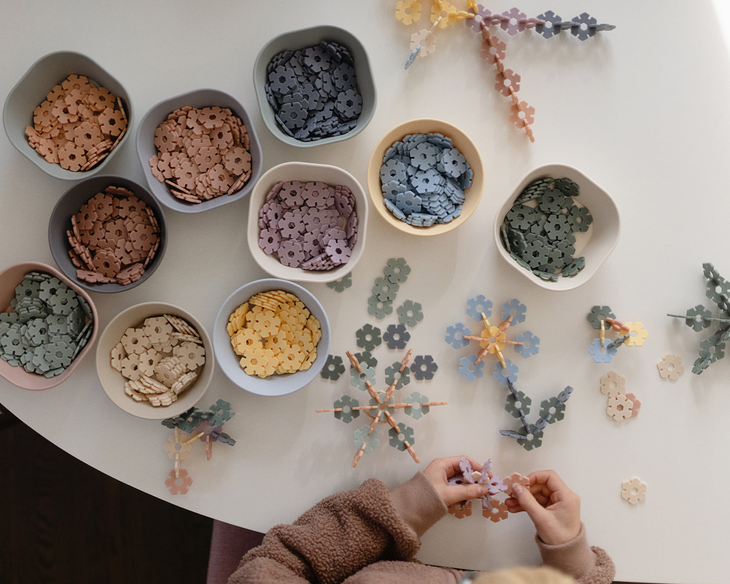 A child’s hands assemble colorful, flower-shaped building pieces on a white table. Several pastel bowls filled with similar pieces are arranged on the table, along with completed star and flower-shaped constructions.