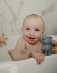 Smiling baby in a tub with mushie Mold Free Bath Play Set and floating toys, easy to clean.