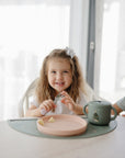 Girl with a bow sits at table with food on mushie Silicone Placemat and mushie cup.