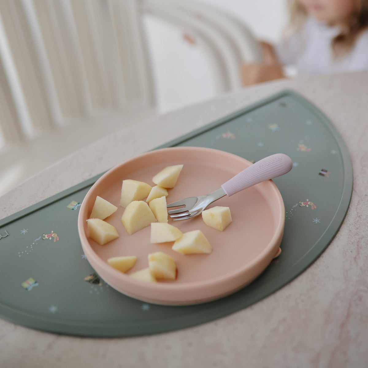 Pink plate with apples and fork on mushie Silicone Placemat; child blurred in the background.