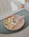 Pink plate with apples and fork on mushie Silicone Placemat; child blurred in the background.