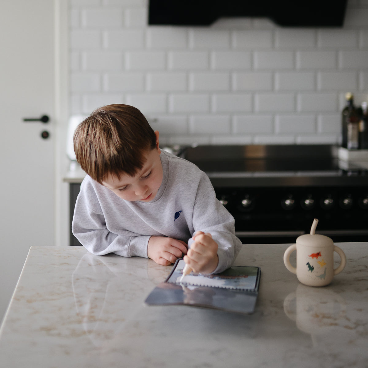 A boy writes at a counter with a Mushie Silicone Training Cup + Straw and a mug nearby.