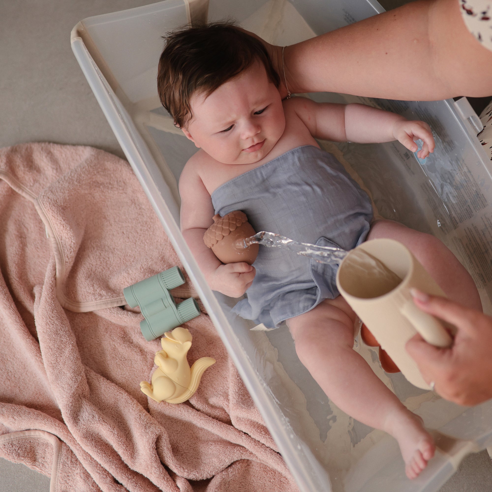 An adult gently bathes a baby in a small plastic tub by pouring water with the Mushie Bath Rinse Cup. The baby, in gray cloth, is holding a toy and surrounded by a pink towel, yellow squirrel toy, and green binoculars.