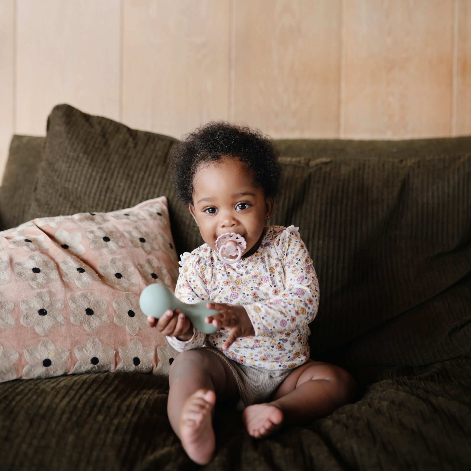 A baby with a FRIGG Andersen Fairytale Silicone Pacifier sits on a dark green couch, holding a light blue rattle. A patterned pillow is beside the baby and a light wooden wall is behind. The pacifier by FRIGG is made in Denmark from safe silicone.