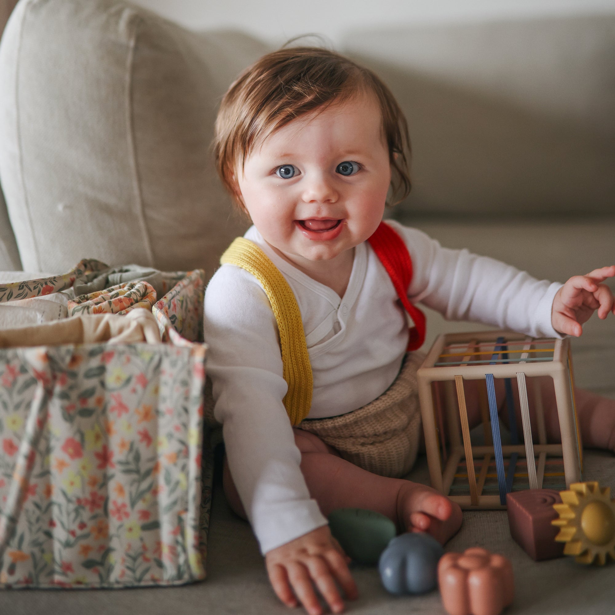 Lifestyle image of a baby sitting on a couch with the Elastic Shape Sorting toy and a Pastel Blooms Diaper Caddy around, wearing a white shirt, yellow suspenders, and a red bib