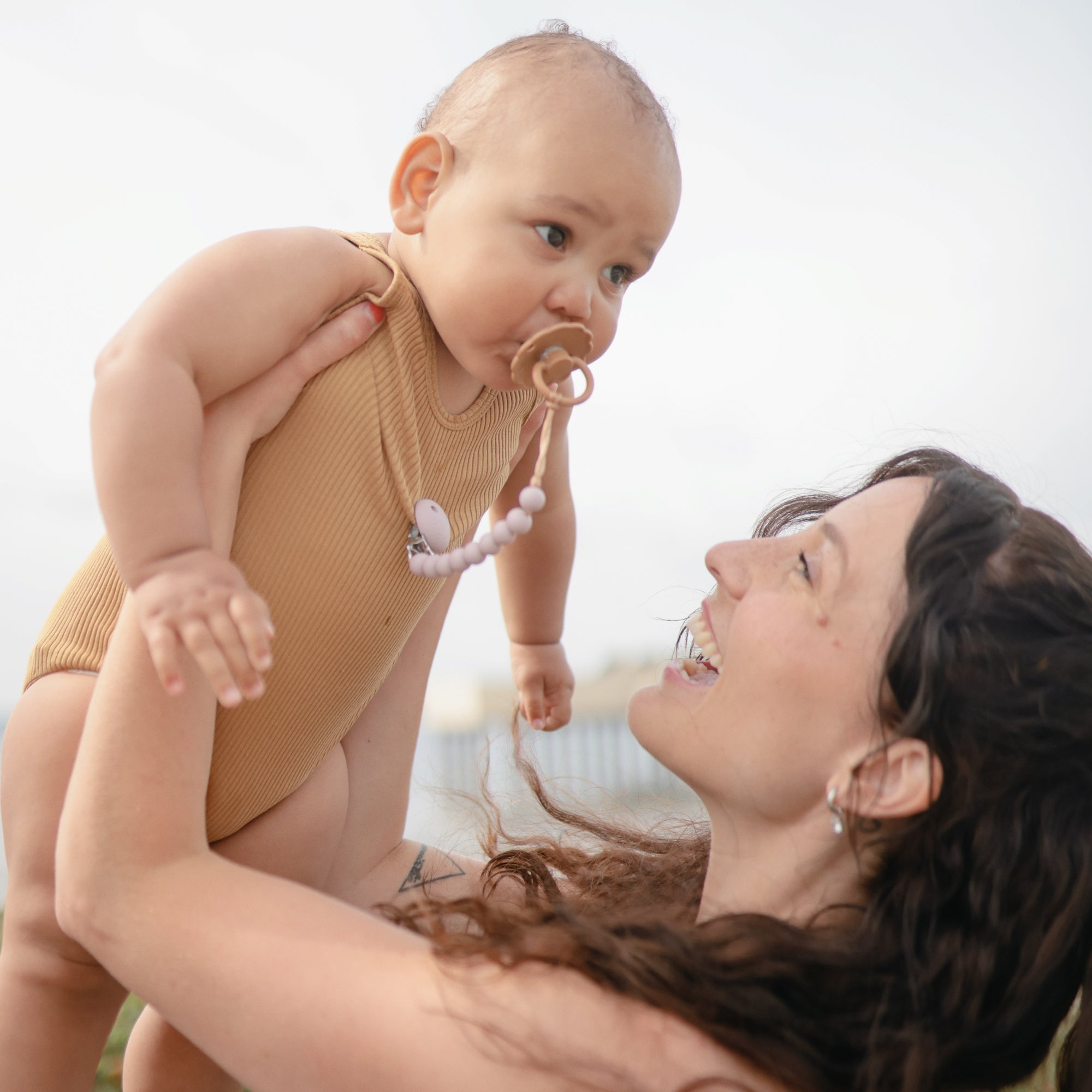 A woman with long brown hair smiles as she lifts a baby in a beige onesie, soothed with the FRIGG Baby's First Pacifier Floral Heart (Blush) 4-Pack by FRIGG. They are outdoors, and the softly blurred background adds warmth to the scene.