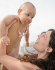 A woman with long brown hair smiles as she lifts a baby in a beige onesie, soothed with the FRIGG Baby's First Pacifier Floral Heart (Blush) 4-Pack by FRIGG. They are outdoors, and the softly blurred background adds warmth to the scene.