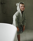 Smiling child holds mushie Suction Spinner Toys by the tub, wearing a bear-eared bathrobe.