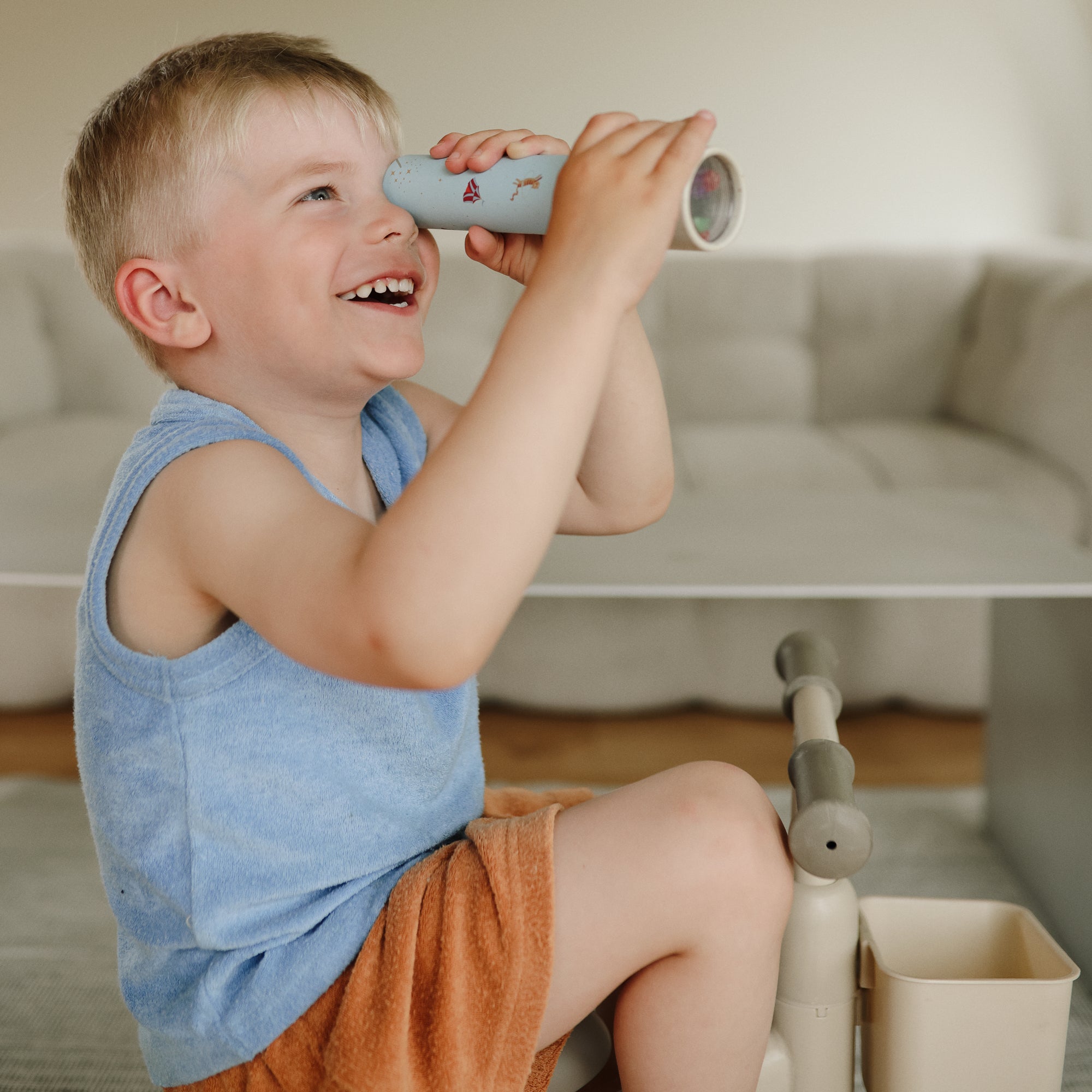A smiling boy explores with a mushie Kaleidoscope while sitting on a cushioned chair.
