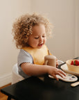 A toddler wearing a mushie Silicone Baby Bib sits at a table with a bowl of strawberries.