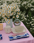 A table with daisies, dishes, mushie Dinnerware Cups, and napkins on a pink cloth outdoors.