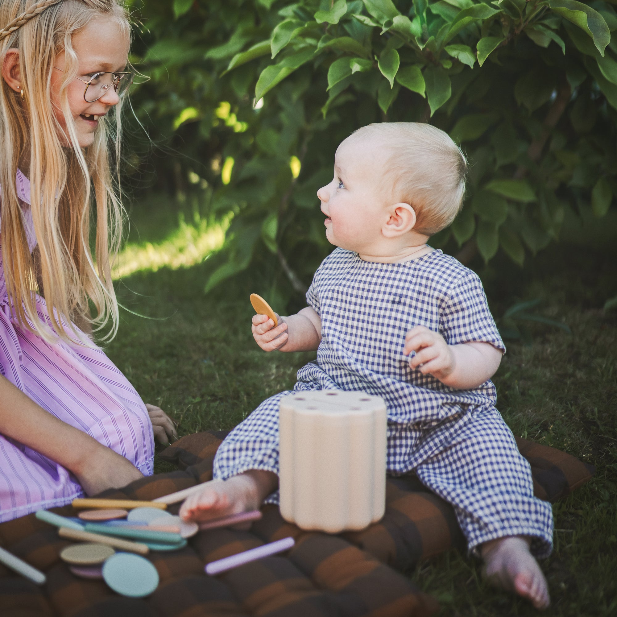 Two children sitting on a blanket outdoors with the mushie Coin and Tube Sorting toy. 