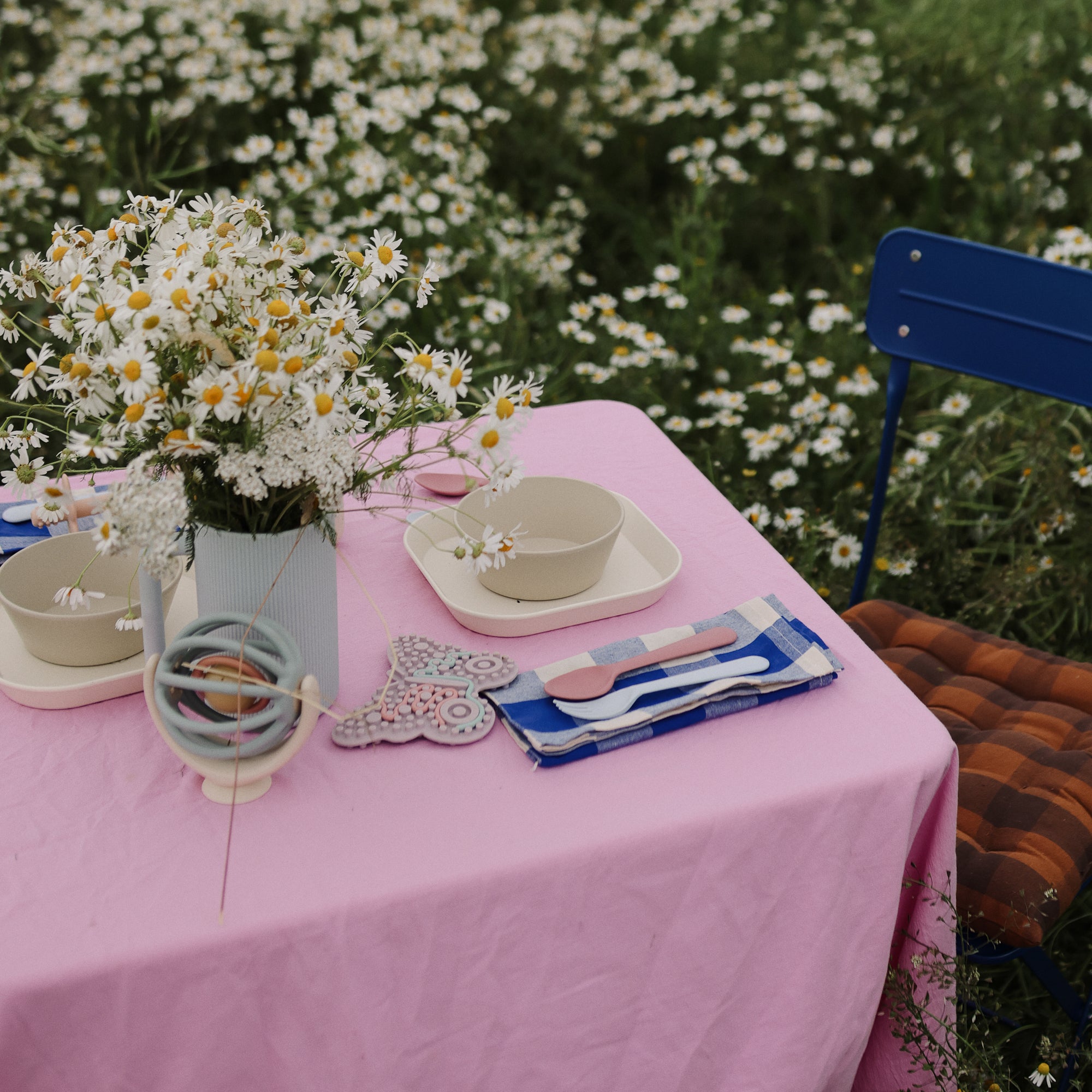 A table with a pink cloth, daisies, dishes, and a Mushie Bath Rinse Cup in a garden setting.