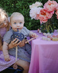 A baby with a mushie Linen Pacifier Clip 2-Pack uses a sippy cup on a girl’s lap outside.