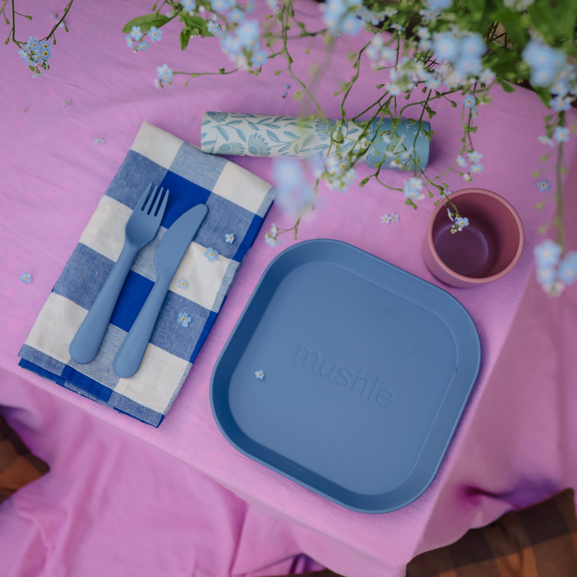 Mushie Dinnerware Cups with blue plate, cutlery on pink floral tablecloth and checkered napkin.