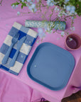 Mushie Dinnerware Cups with blue plate, cutlery on pink floral tablecloth and checkered napkin.