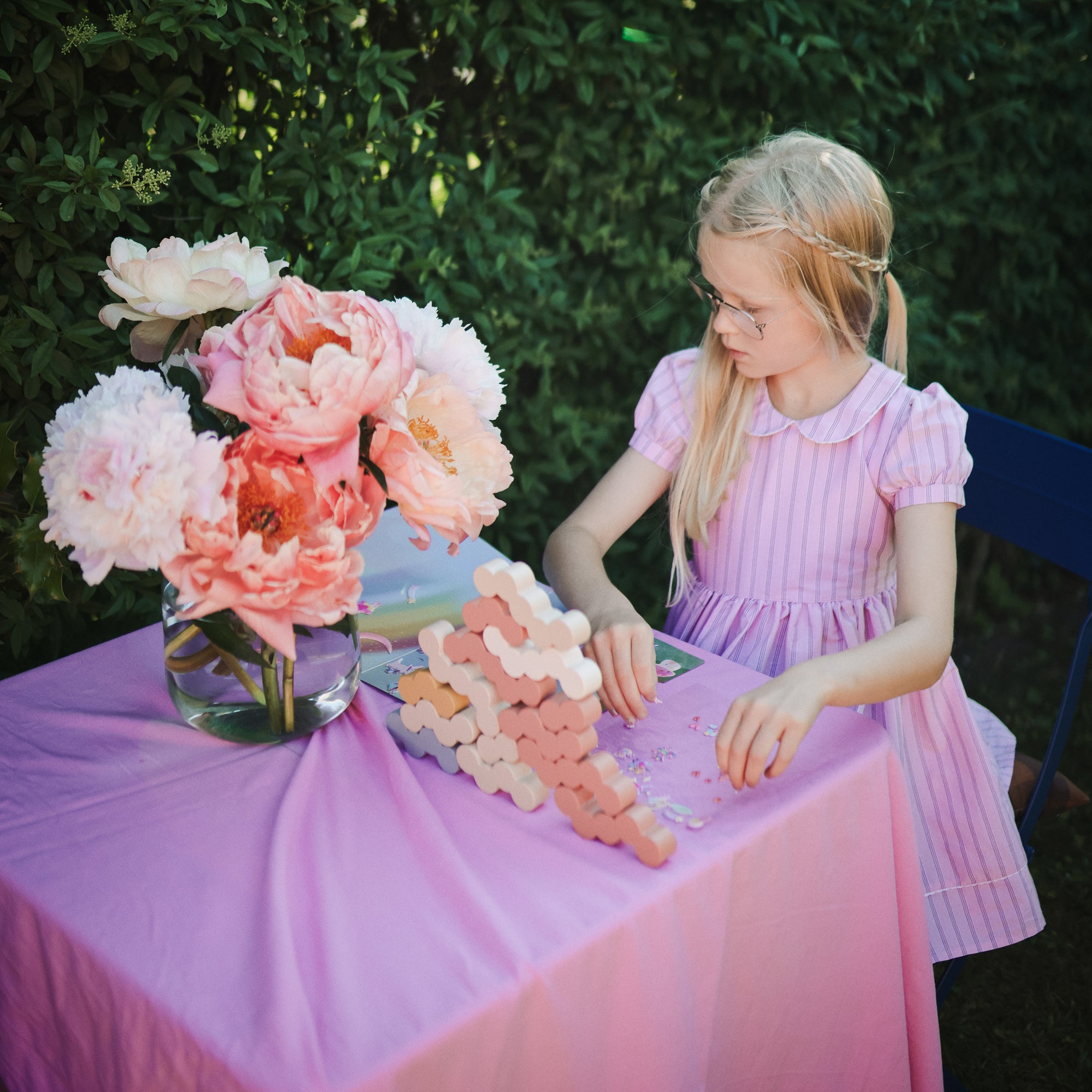A girl sits at a small table outdoors, playing with mushie Wooden Balance Blocks and Fairies Reusable Sticker Set. 