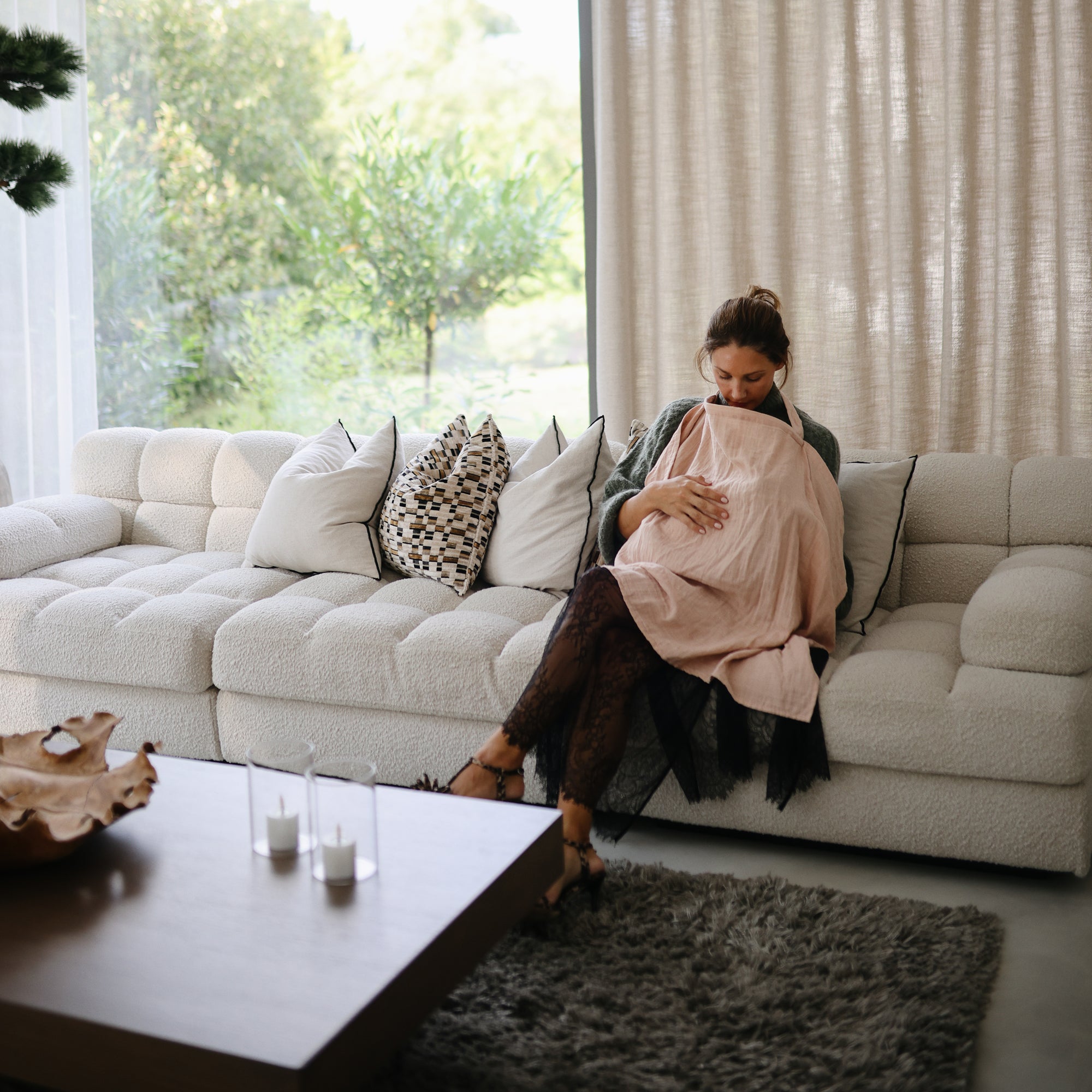 A woman sits on a white couch in a bright, modern living room, breastfeeding her baby under the mushie Muslin Nursing Cover. Surrounded by cushions, she looks down at her baby near a coffee table and large windows.