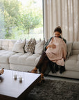 A woman sits on a white couch in a bright, modern living room, breastfeeding her baby under the mushie Muslin Nursing Cover. Surrounded by cushions, she looks down at her baby near a coffee table and large windows.