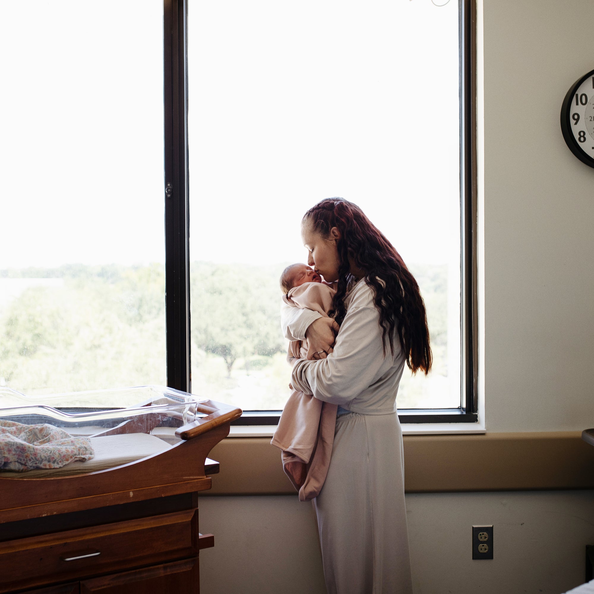 A woman in a light robe stands by a large window, gently holding and kissing her newborn wrapped in the Mushie Ribbed Baby Blanket. A hospital bassinet sits nearby as the clock shows 10:09 with trees visible outside.