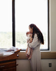 A woman in a light robe stands by a large window, gently holding and kissing her newborn wrapped in the Mushie Ribbed Baby Blanket. A hospital bassinet sits nearby as the clock shows 10:09 with trees visible outside.