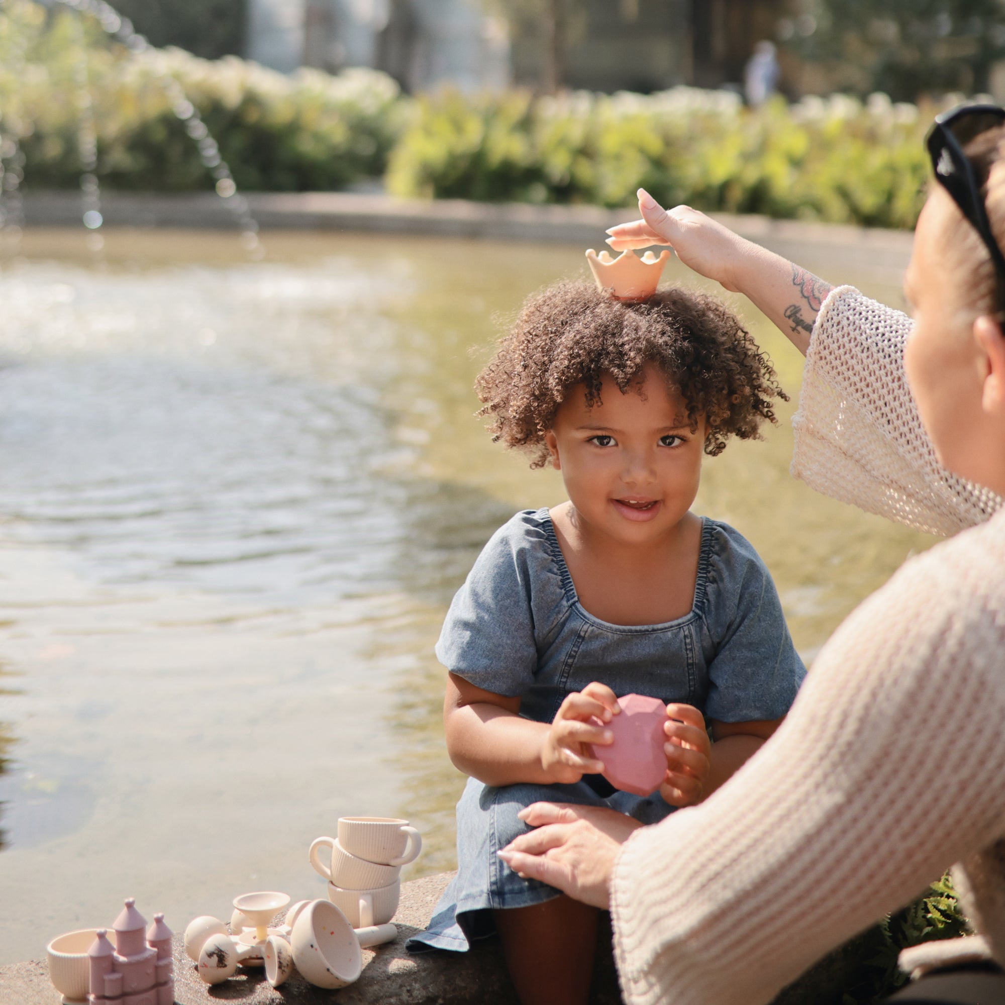 A woman crowns a smiling girl near mushie Mold Free Bath Play Set, easy to clean and safe.