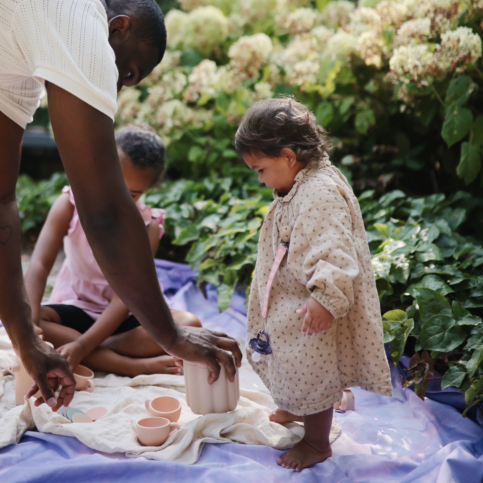 An adult and two children enjoy an outdoor tea party on a blanket among green plants, as FRIGG Daisy Silicone Pacifier 2-Pack by FRIGG rests nearby while the kids play.