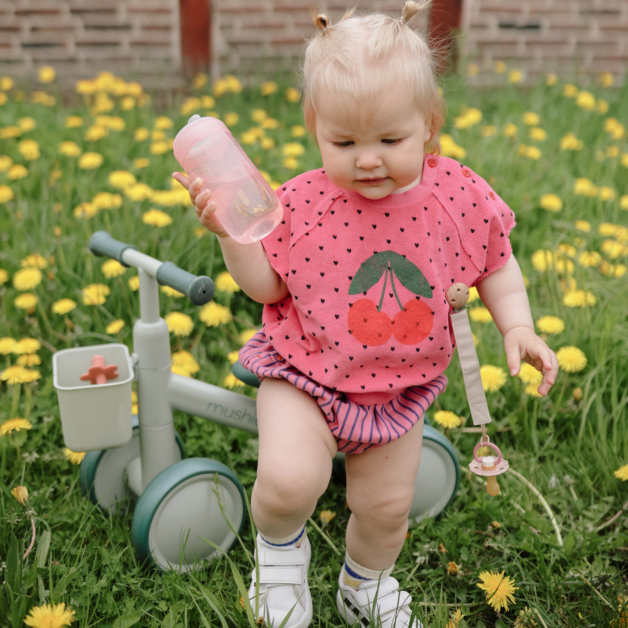 Toddler with mushie Linen Pacifier Clip 2-Pack sits on a balance bike among dandelions.