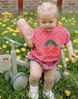 Toddler in pink with bottle on a mushie Mini Toddler Bike, surrounded by dandelions.