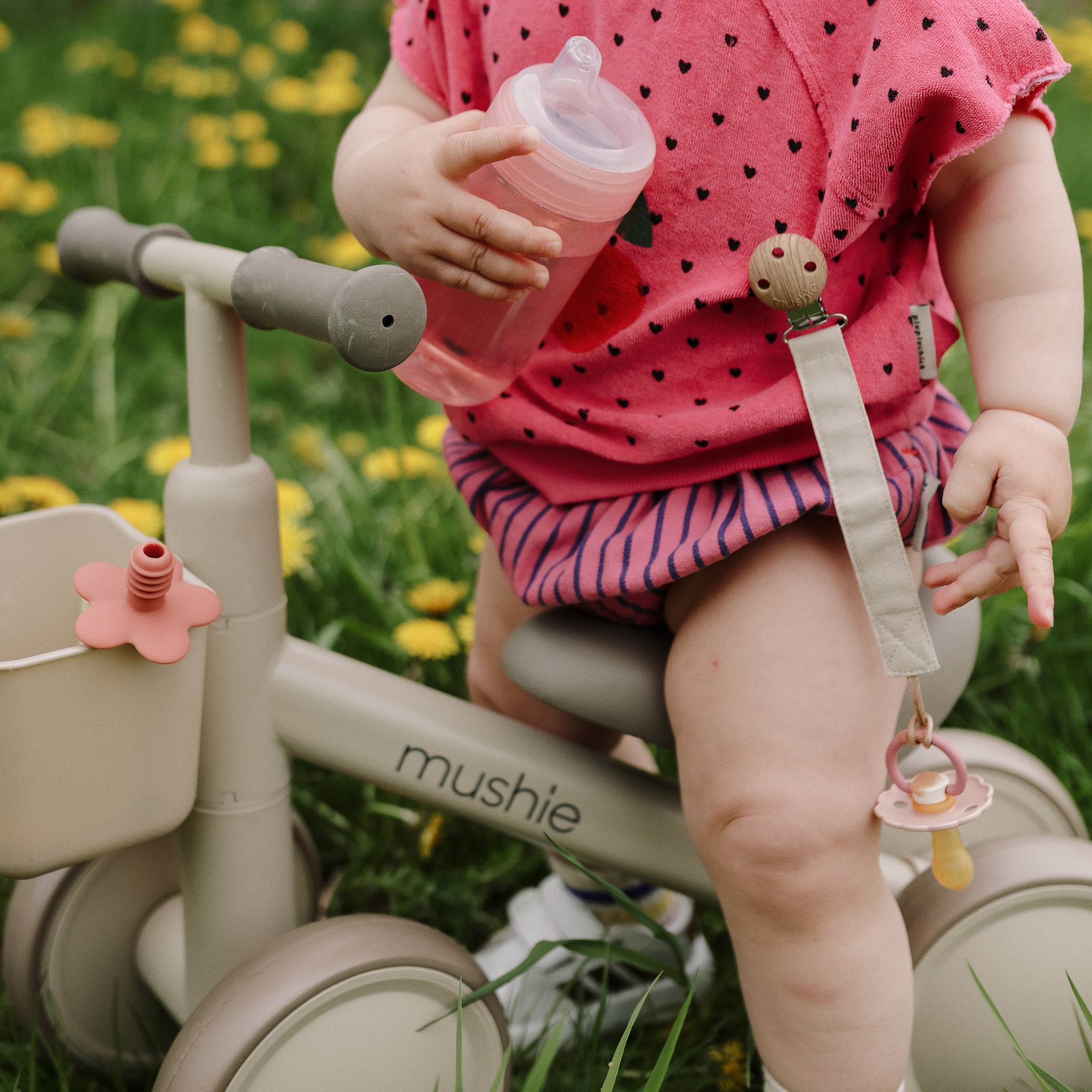 A toddler rides a mushie Mini Toddler Bike, holding a bottle with a pacifier clipped on.