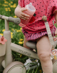 A toddler rides a mushie Mini Toddler Bike, holding a bottle with a pacifier clipped on.