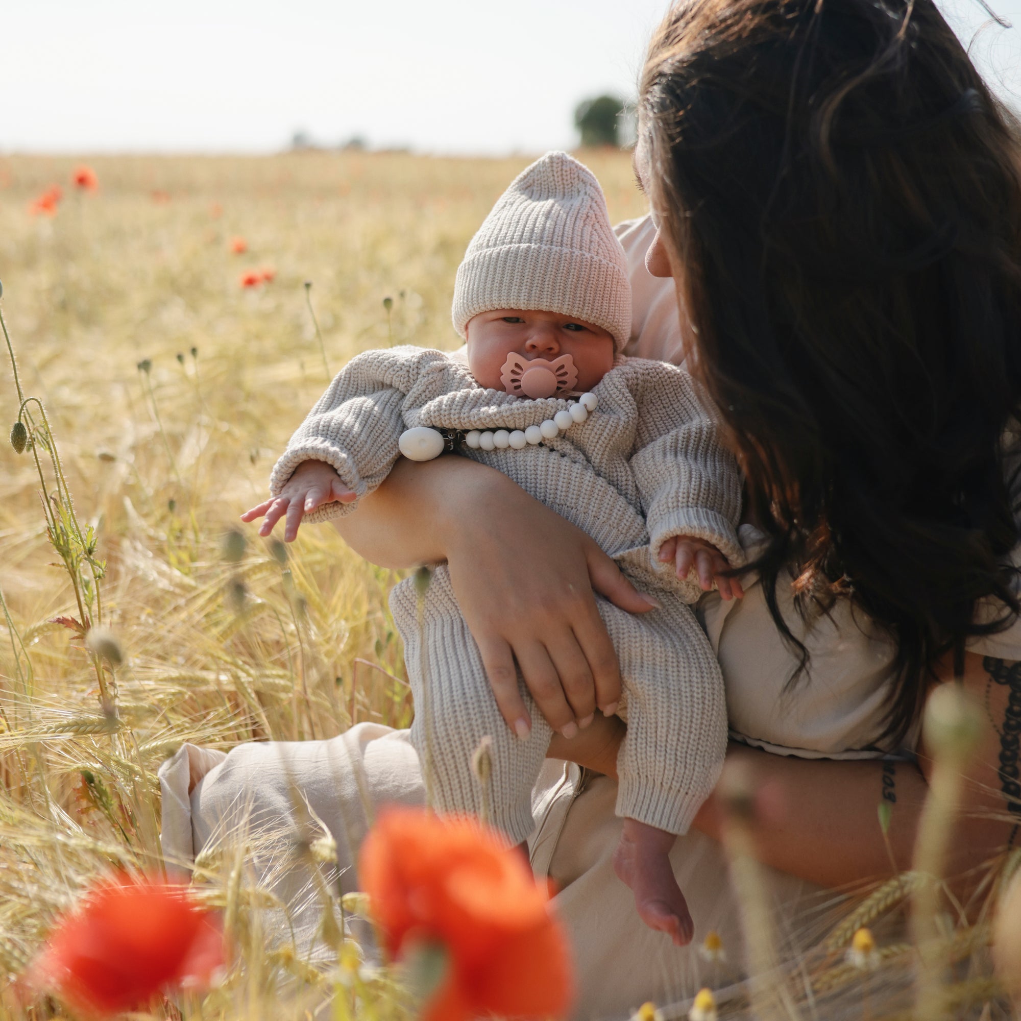 A woman holds a baby in a knitted outfit and hat among tall grass and red poppies. The calm baby gently sucks on the FRIGG Lucky Symmetrical Silicone Pacifier (0-6 Months), supporting oral development.