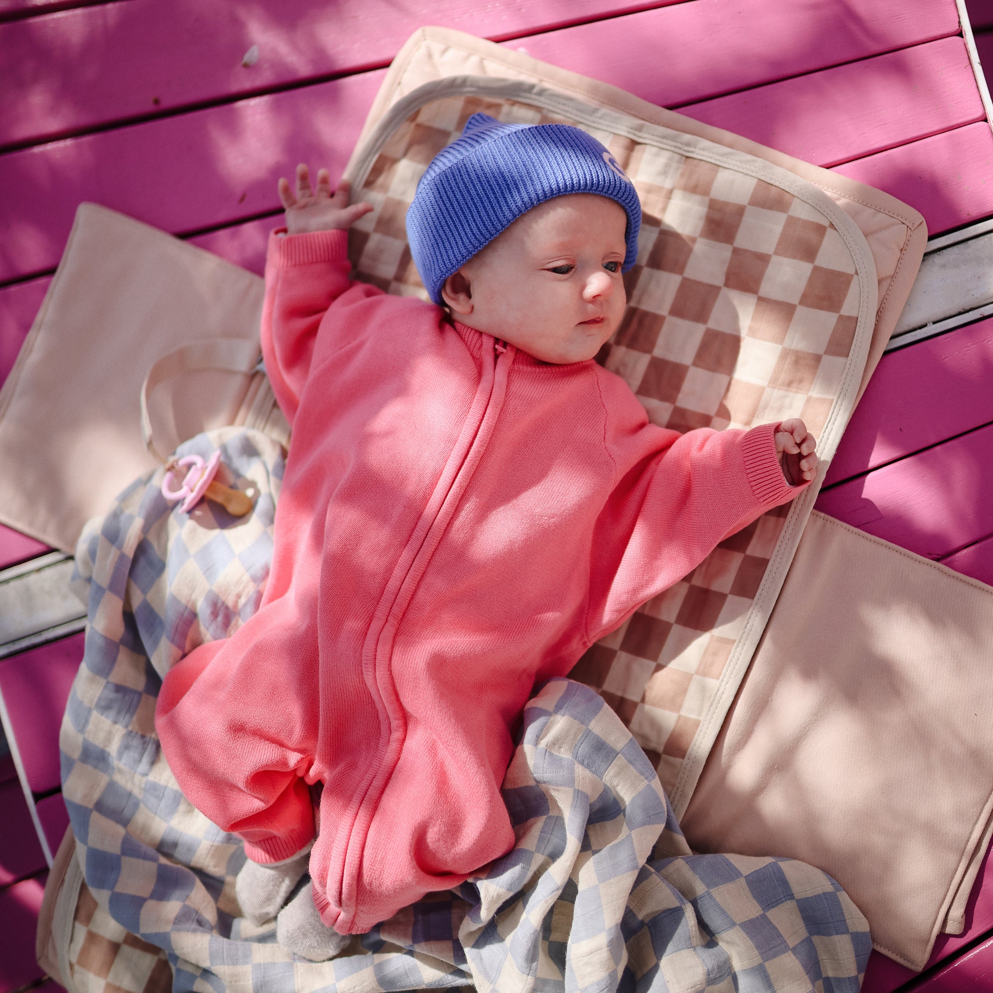 Baby in pink onesie and blue hat on Mushie Changing Pad Liners, with a checkered blanket.