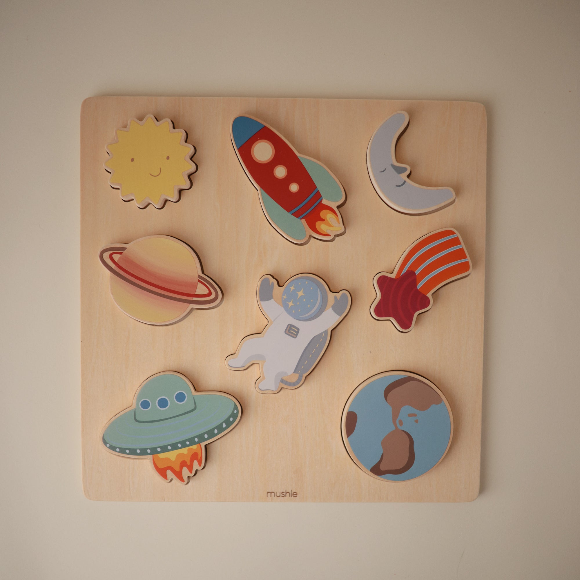 A wooden puzzle featuring colorful pieces: the sun, a rocket, the moon, Saturn, an astronaut, a comet, a flying saucer, and planet Earth, all arranged on a square wooden board.