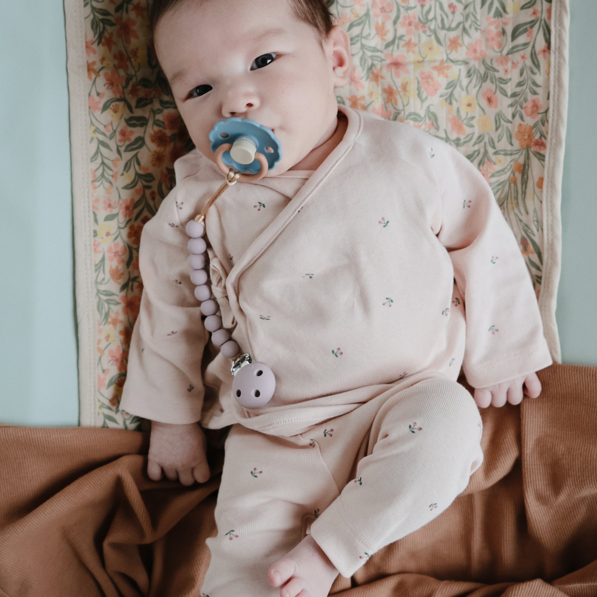 A baby in pale pink floral pajamas lies on a floral mat, holding a pacifier with a beaded clip, surrounded by the Mushie Ribbed Baby Blanket in warm brown.