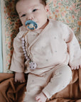 A baby in pale pink floral pajamas lies on a floral mat, holding a pacifier with a beaded clip, surrounded by the Mushie Ribbed Baby Blanket in warm brown.