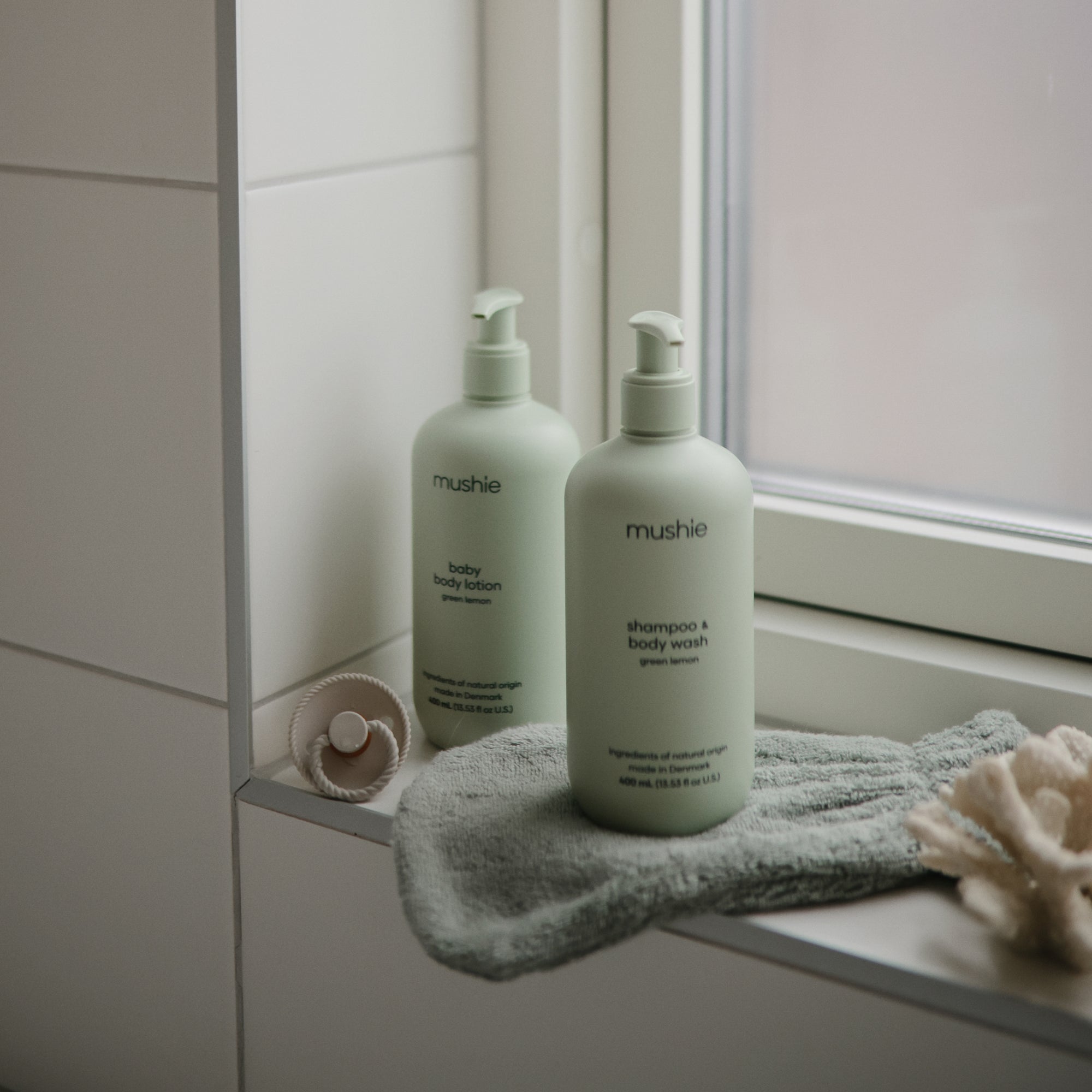 Two green pump bottles labeled mushie baby body lotion and shampoo &amp; body wash sit on a bathroom windowsill with a green washcloth, a pacifier, and a cream-colored sponge nearby.