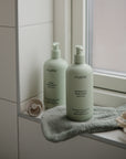 Two light green bottles of Mushie baby body lotion and shampoo & body wash sit on a windowsill with baby essentials. They pair perfectly with Mushie's FRIGG Rope Natural Rubber Pacifier 2-Pack.