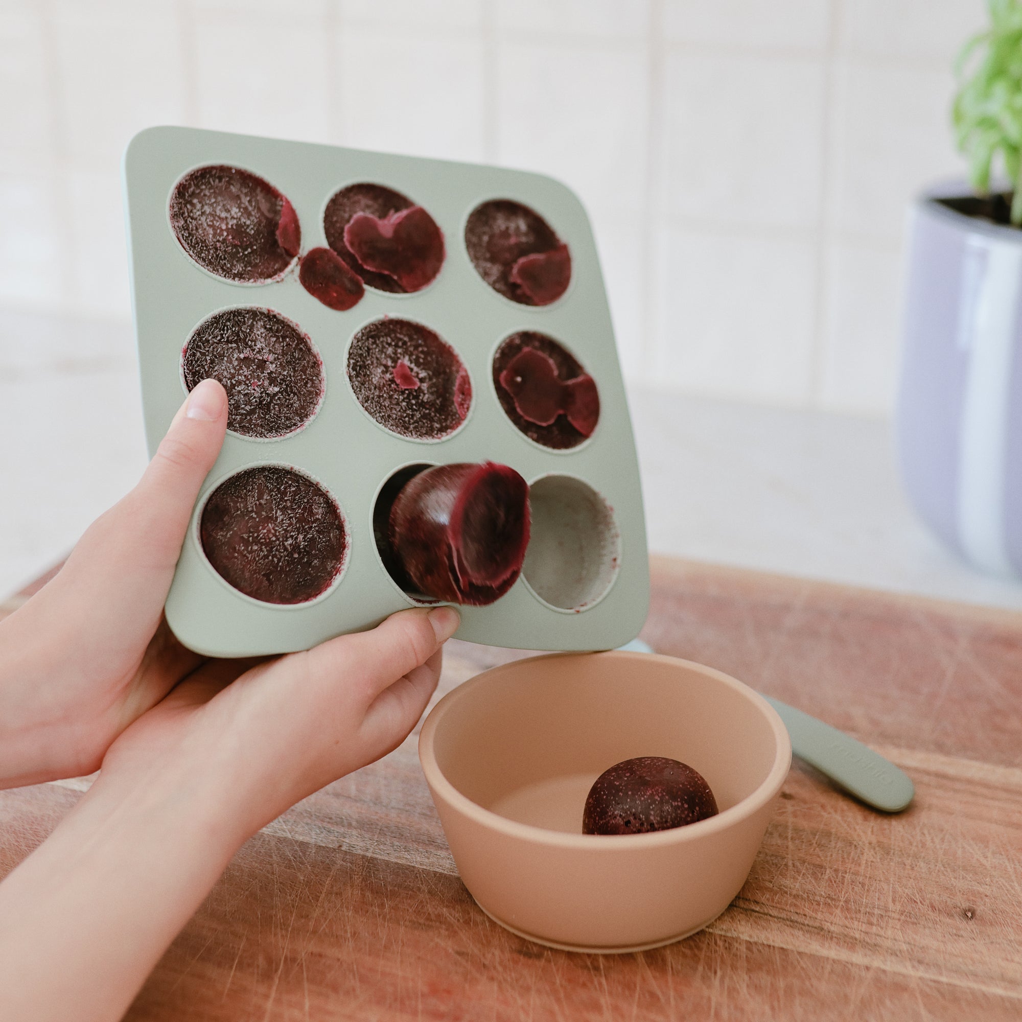 Hands pop frozen fruit puree from a Mushie Baby Food Freezer Tray into a beige bowl on wood.