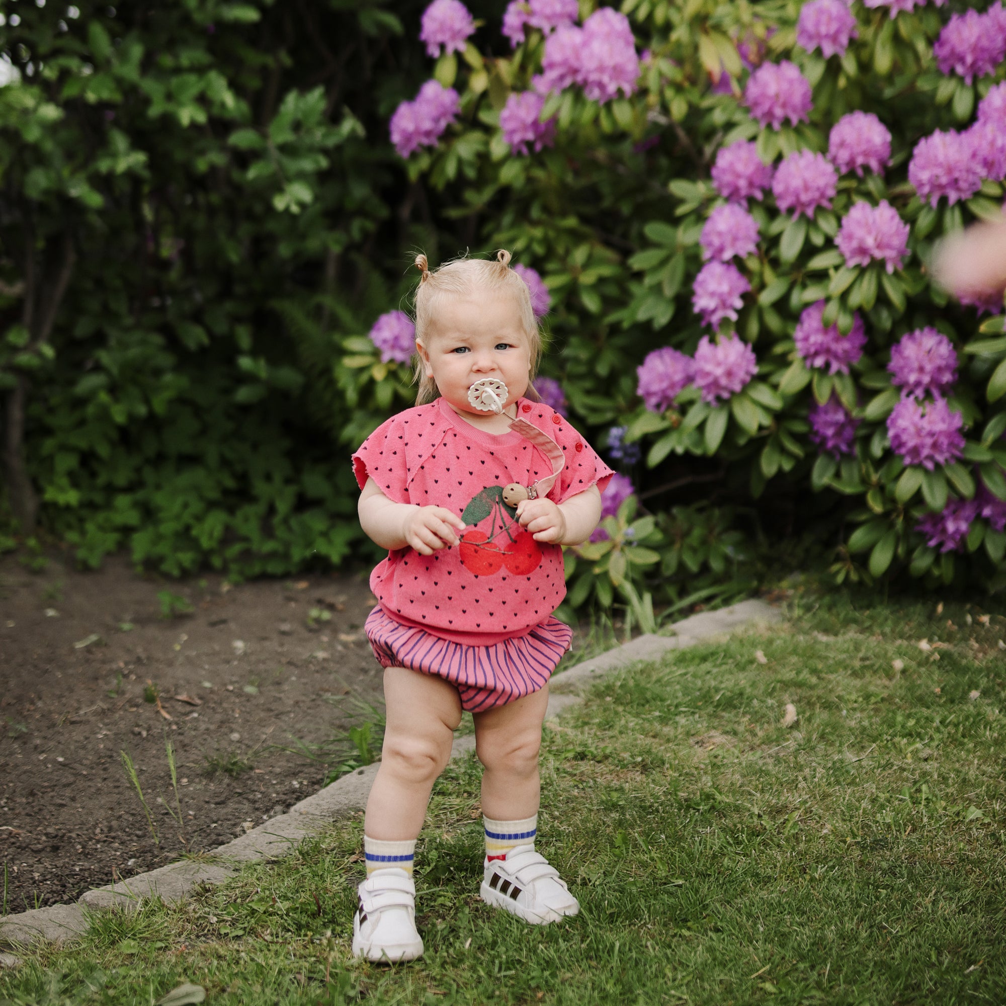A toddler in pink stands near flowers, holding a dandelion and a FRIGG silicone pacifier.