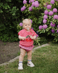 A toddler in pink stands near flowers, holding a dandelion and a FRIGG silicone pacifier.