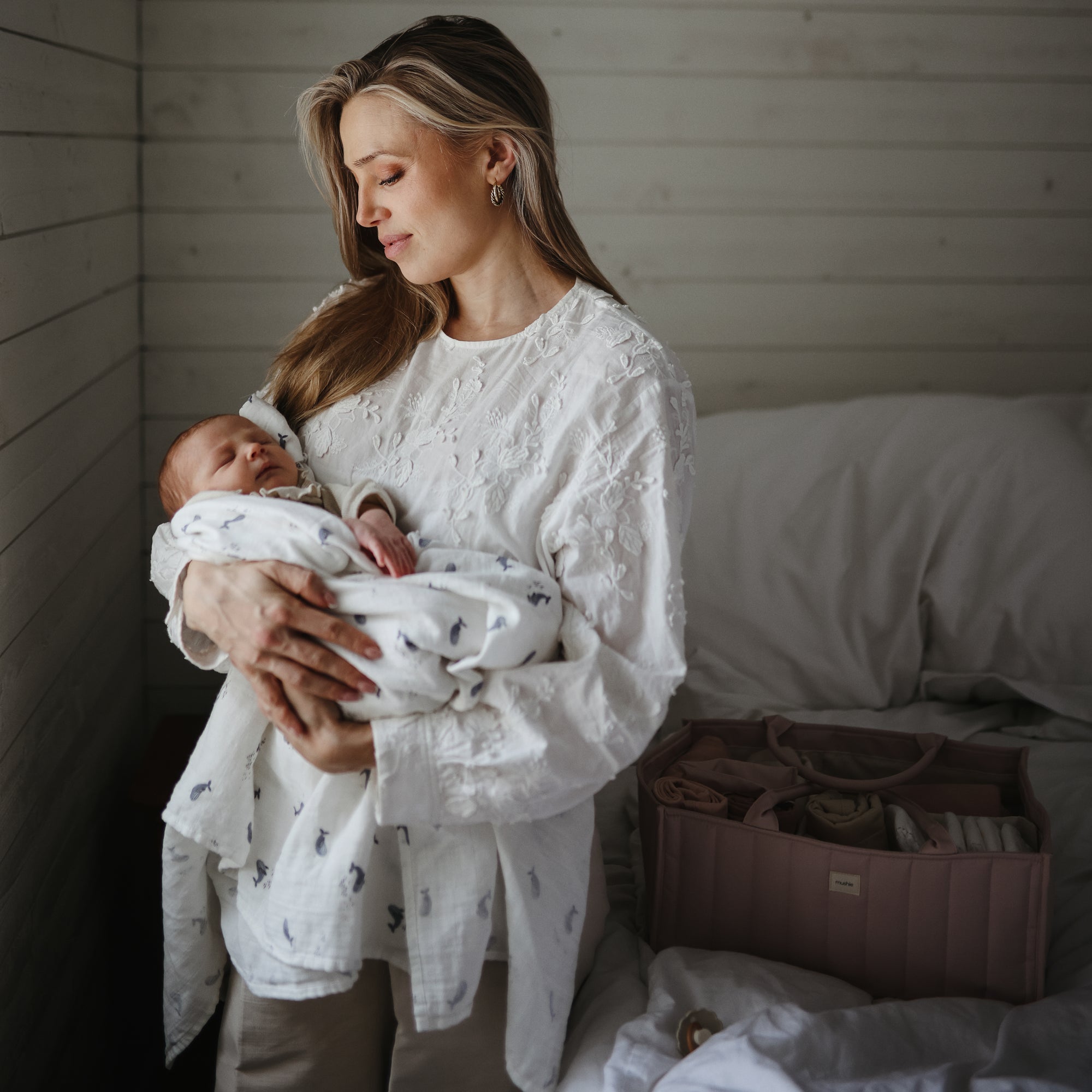 A woman in a white blouse gently cradles her newborn wrapped in a mushie Organic Cotton Muslin Swaddle Blanket, in a softly lit bedroom featuring white wooden walls and a pink diaper bag on the bed.