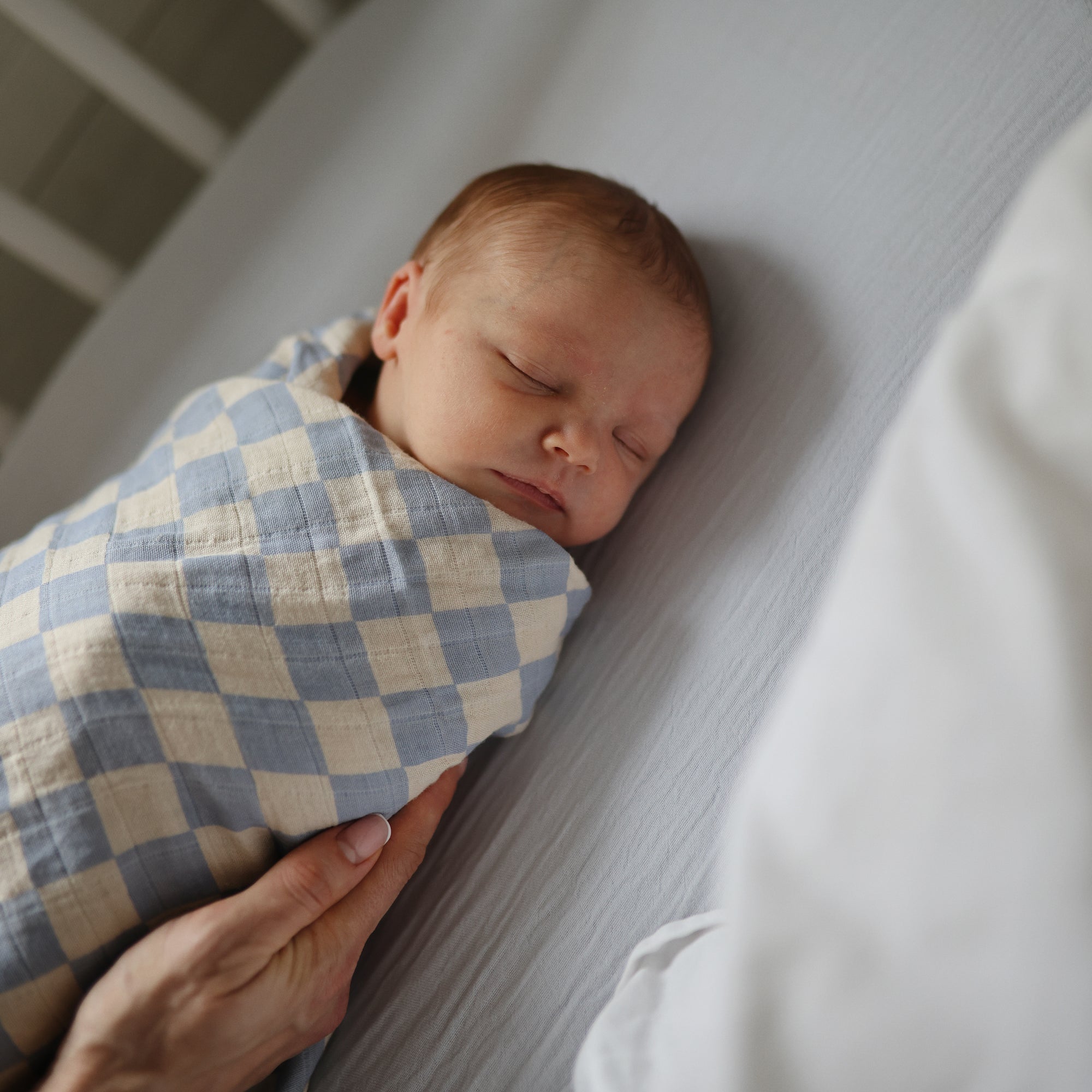A newborn sleeps peacefully, wrapped in a mushie Organic Cotton Muslin Swaddle Blanket in blue and white checks. An adult hand gently comforts the baby, who lies on a soft white crib sheet.