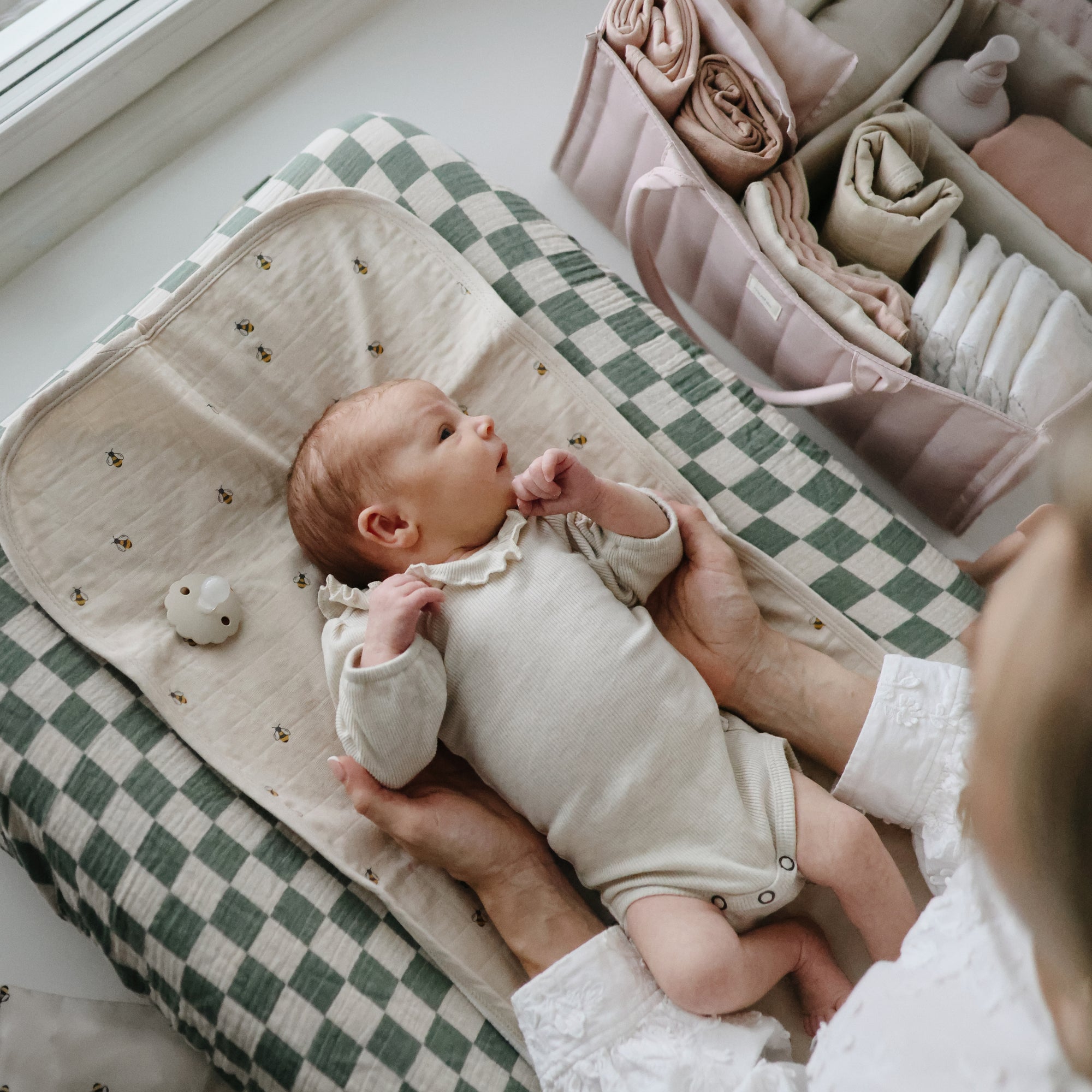 A baby in a cream onesie lies on the mushie Extra Soft Muslin Changing Pad Cover with a checkered pattern. An adult holds the baby gently, while pastel baby clothes and accessories are neatly arranged nearby.