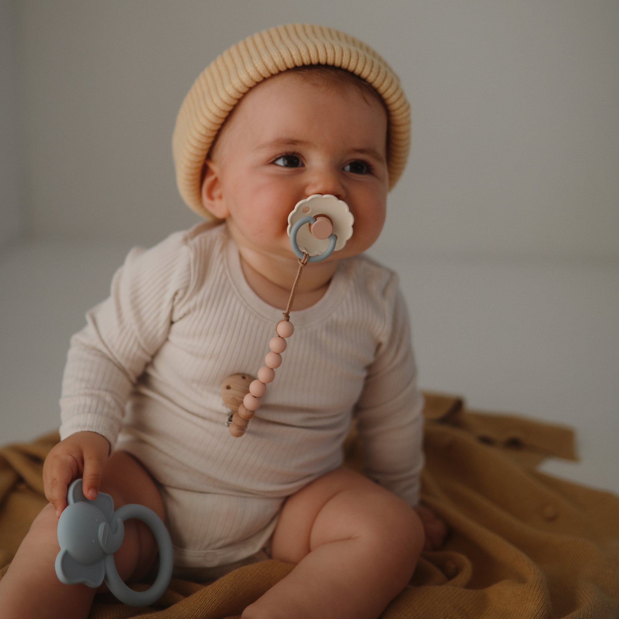 A baby in a beige knit hat sits on a mustard blanket, holding a blue teether and using the FRIGG Daisy Silicone Pacifier 2-Pack by FRIGG with a beaded clip for added comfort.
