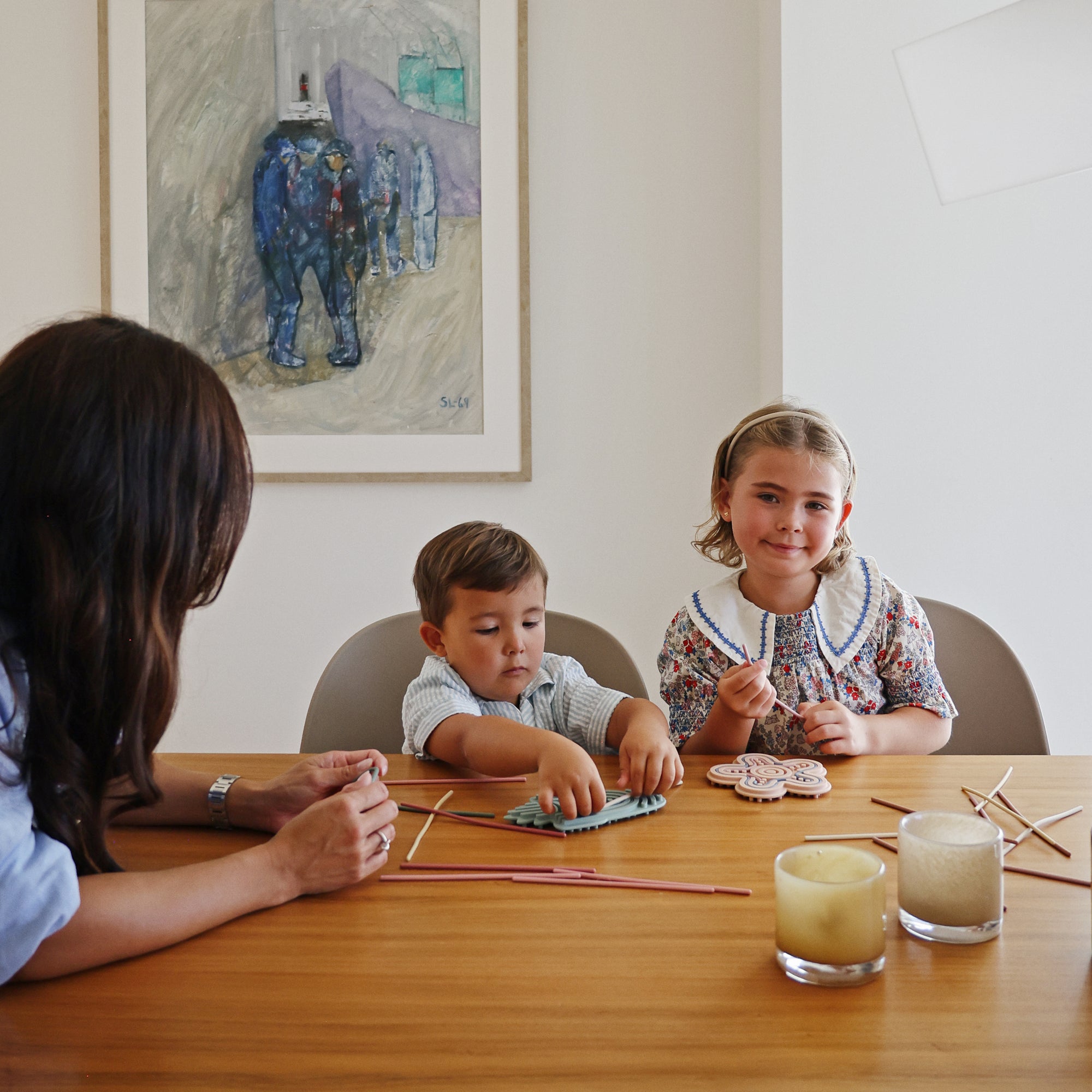 A woman and two kids craft at the table with candles, art, and a mushie Silicone Sensory Toy.