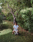 A young boy rides his mushie Mini Toddler Bike on grass under leafy trees in a garden.
