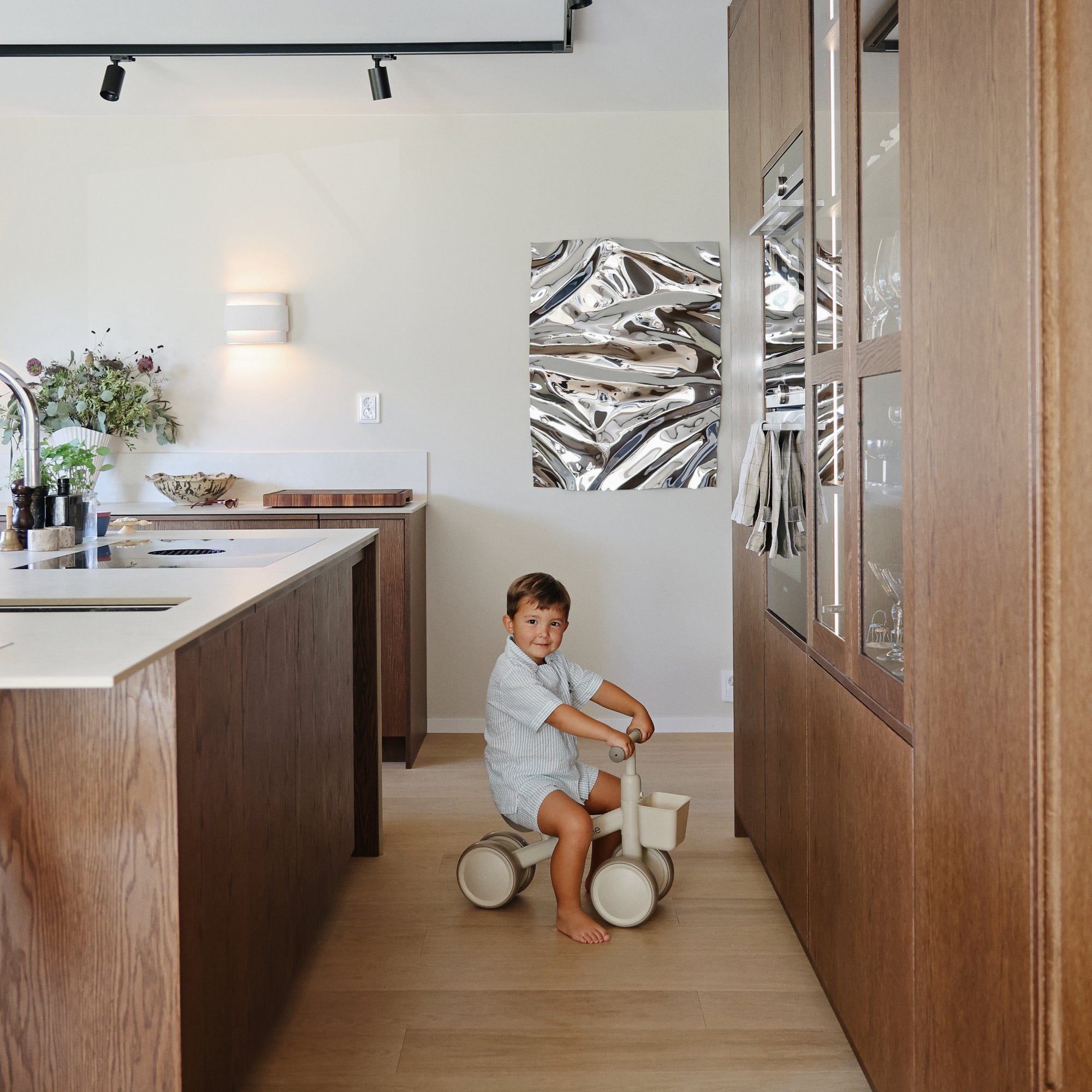 A toddler rides a mushie Mini Toddler Bike in a modern kitchen with wood and metal decor.
