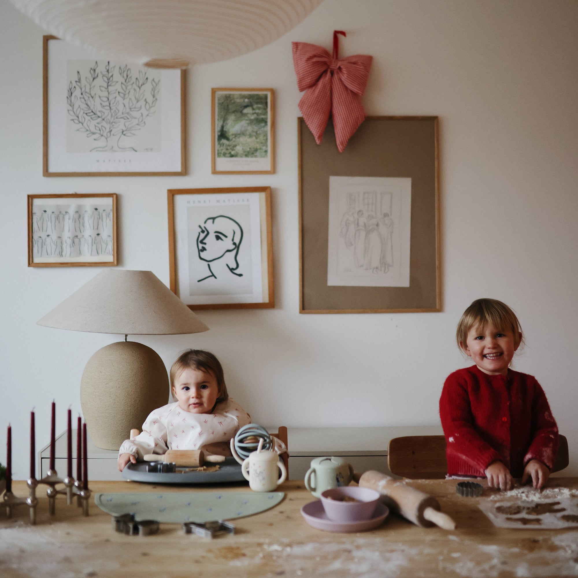 Two young children smile as they make cookies at a wooden table covered in baking supplies, flour, and a mushie Round Dinnerware Bowl. Framed art and a red bow decorate the wall, adding to the warm, cozy atmosphere.