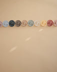 Five FRIGG Moon Natural Rubber Pacifier 2-Packs from FRIGG, featuring assorted colors, are arranged in a line on a beige surface under soft lighting with gentle shadows—an essential for any baby.